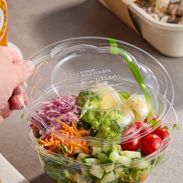 A clear compostable dome lid being placed on a deli bowl filled with salad ingredients.