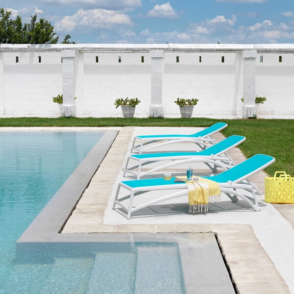 A white adjustable stackable resin chaise lounge with a light blue fabric seat, positioned beside a pool.
