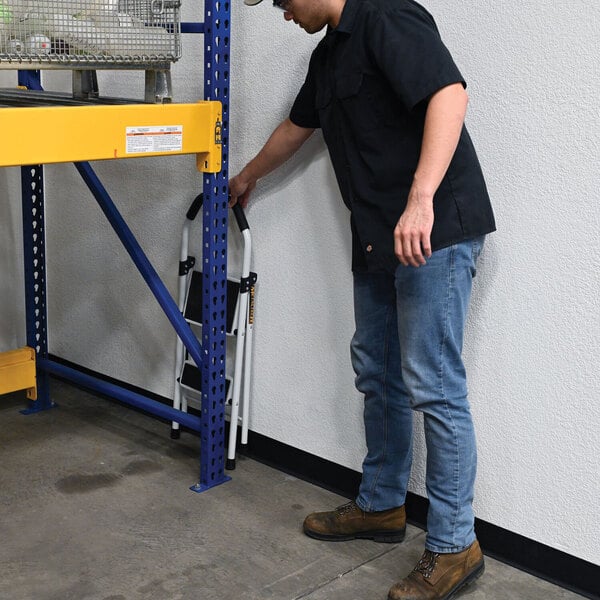 A person placing a folded Vestil steel 2-step fold-up step ladder against a wall in an industrial setting.