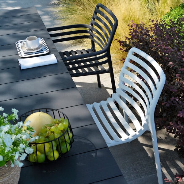 A black resin stackable outdoor arm chair with a slatted design, placed next to a table in an outdoor setting.
