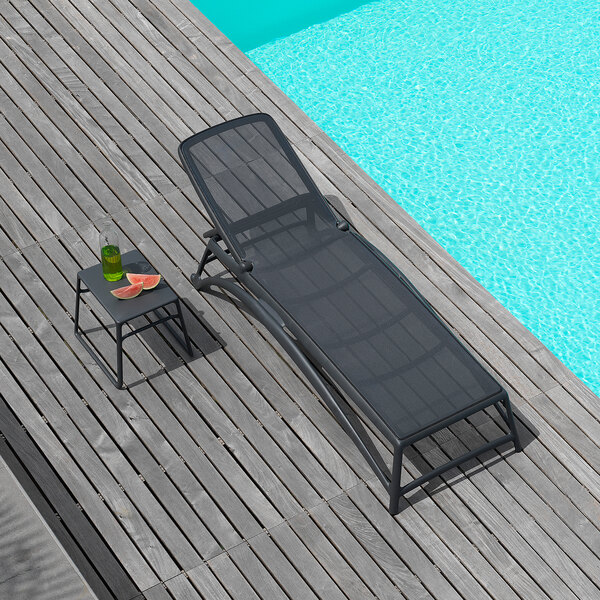 A black adjustable resin chaise lounge with a matching fabric seat, placed on a wooden deck beside a pool.