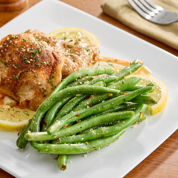 A plate of cooked whole green beans served with seasoned chicken and lemon slices.