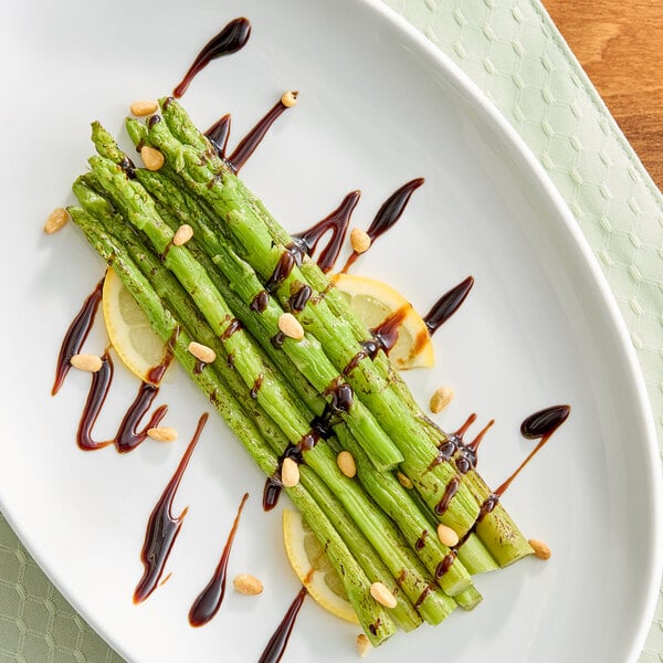 A plate of cooked asparagus spears garnished with pine nuts, lemon slices, and a drizzle of balsamic glaze.