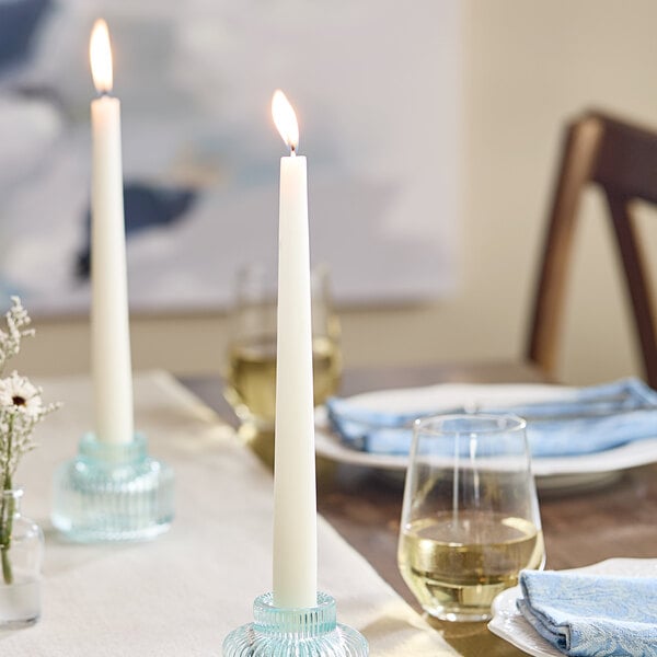A white 10-inch taper candle burning in a glass holder on a set dining table.