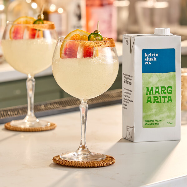 A 32-ounce carton of Kelvin Slush Co. Organic Frozen Margarita Cocktail Mix next to a glass of margarita garnished with fruit and a chili pepper.
