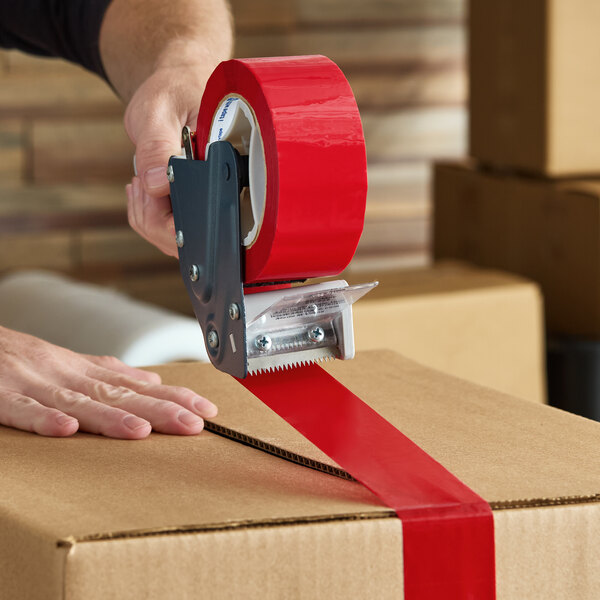 A roll of red acrylic carton sealing tape being applied to a cardboard box with a tape dispenser.