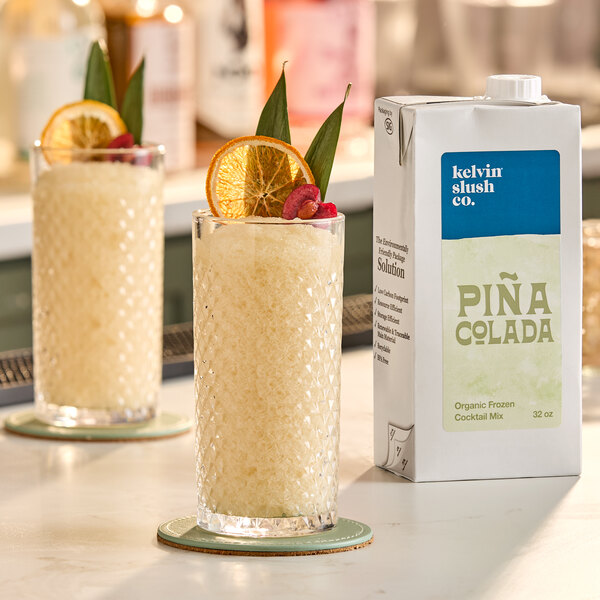 A 32-ounce carton of Kelvin Slush Co. Organic Frozen Piña Colada Cocktail Mix is shown next to a glass of frozen piña colada garnished with a dried orange slice, pineapple leaves, and a cherry.