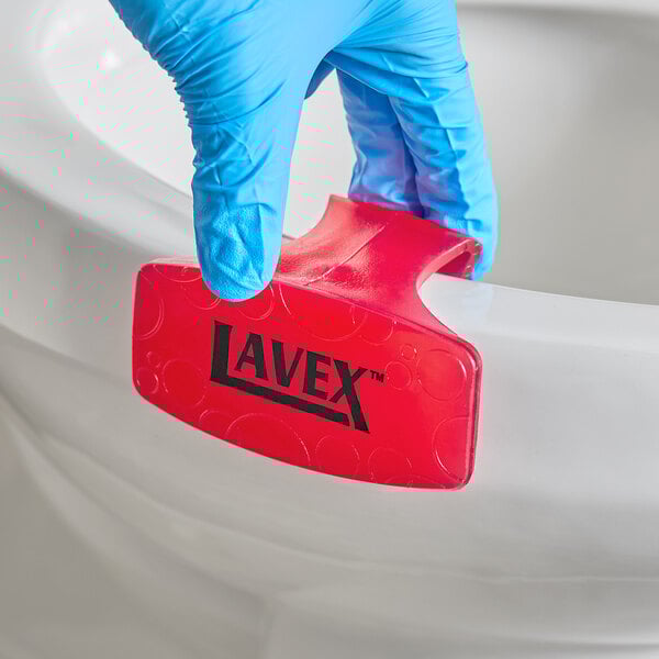 A red Lavex toilet bowl clip being placed on the rim of a toilet by a gloved hand.