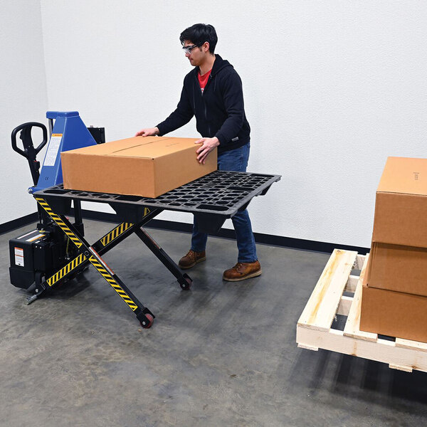 A battery-powered tote lifter with 45-inch by 27-inch forks and a photo eye sensor, shown lifting a large cardboard box in an industrial setting.