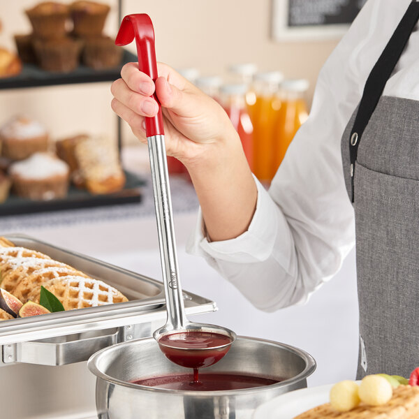 A stainless steel ladle with a red coated handle being used to serve sauce from a metal container.