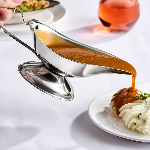 A stainless steel gravy boat pouring brown gravy onto mashed potatoes.