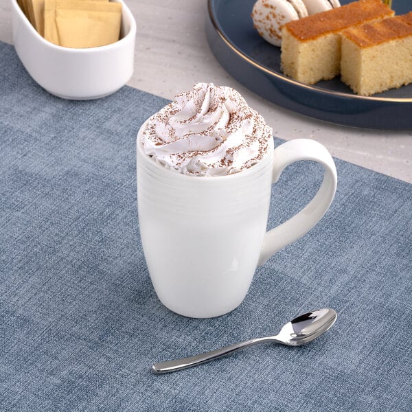 A white embossed porcelain mug filled with a whipped cream-topped beverage, placed on a blue placemat with a spoon nearby.