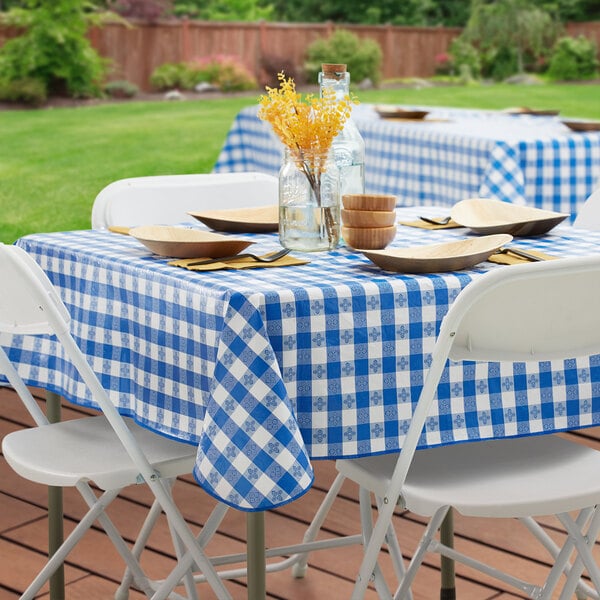 A rectangular table covered with a royal blue and white gingham vinyl tablecloth with a flannel back, set outdoors with plates, bowls, and simple decor.