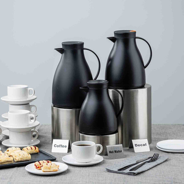 A black plastic coffee carafe with a glass liner, displayed alongside other carafes, cups, and pastries on a table.