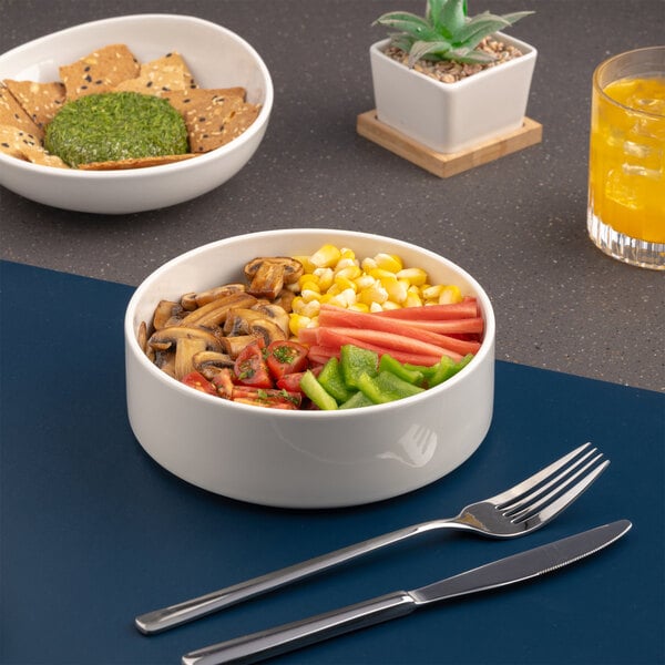 A white porcelain salad bowl filled with assorted vegetables, placed on a table with a fork and knife.