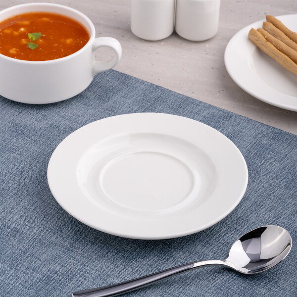 A round, wide rim, warm white porcelain soup bowl saucer placed on a blue placemat.