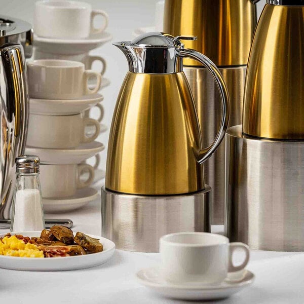 A gold and stainless steel coffee carafe with a handle, surrounded by stacked cups and breakfast items.