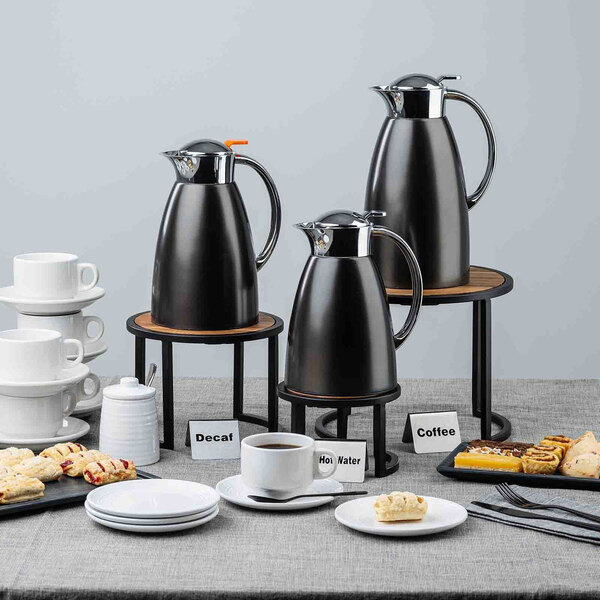 A black stainless steel coffee carafe with a handle and spout, displayed on a table with other breakfast items.