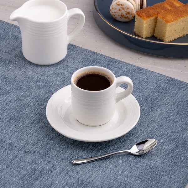 A white embossed porcelain espresso cup with a saucer and spoon, accompanied by a milk jug and desserts in the background.