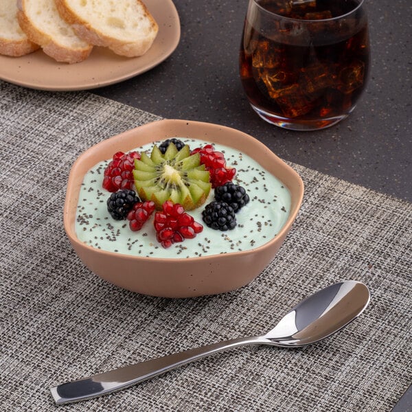 A bowl of Ariane Earth Dark Sand Dots porcelain filled with yogurt, fruit, and a spoon on a table with a glass of brown liquid.