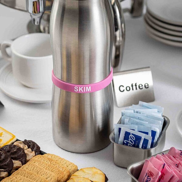 A stainless steel creamer carafe with a pink silicone band labeled 'SKIM' on a table set with coffee service items and assorted cookies.