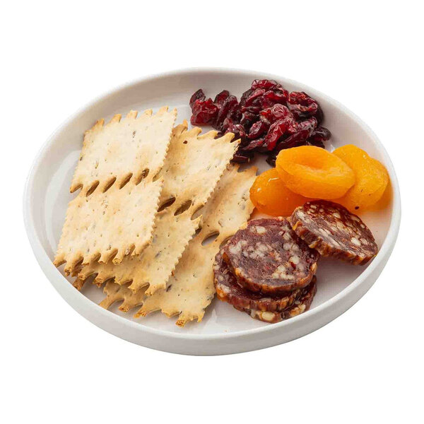 A white round melamine plate containing crackers, dried cranberries, dried apricots, and slices of cured sausage.