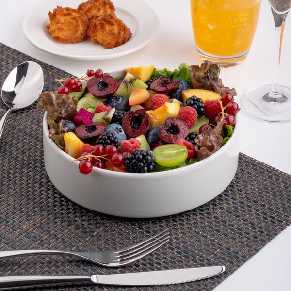 A white porcelain salad bowl filled with assorted fresh fruits, placed on a woven placemat with cutlery nearby.
