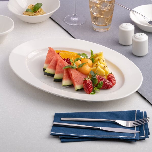 An oval wide rim porcelain platter in warm white, measuring 11 by 7 11/16 inches, displayed with assorted fruit.