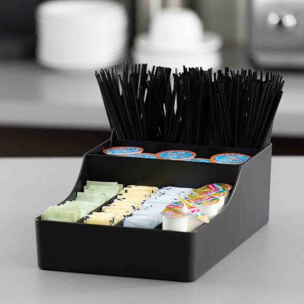 A black plastic coffee condiment organizer with nine compartments holding stirrers, creamers, sugar packets, and other coffee accessories.