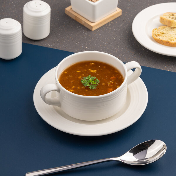 A white embossed porcelain soup bowl with handles, filled with soup and placed on a matching saucer.