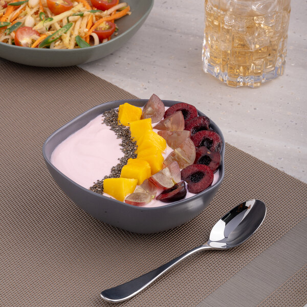 A bowl of fruit and yogurt with a spoon on a table with a glass of liquid.