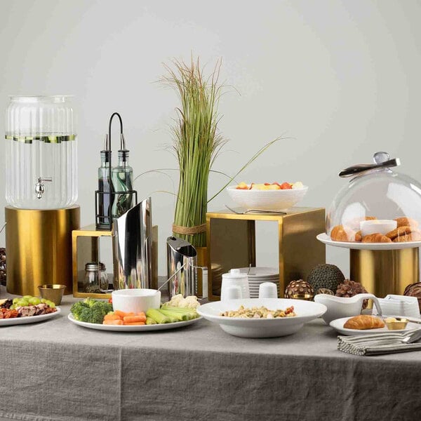 A three-piece round gold stainless steel riser set displayed on a buffet table with various foods and beverages.