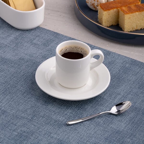 A small white porcelain espresso cup with a matching saucer and a metal spoon on a blue placemat.
