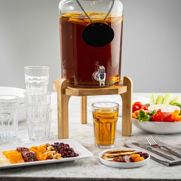 A bamboo beverage dispenser stand holding a glass drink dispenser on a table with glasses and food platters.