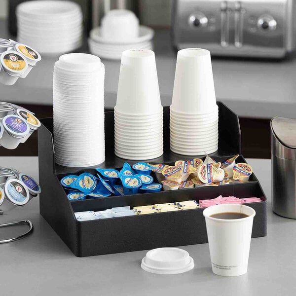 A black plastic coffee condiment organizer with four compartments holding cups, creamers, sugar packets, and stirrers.