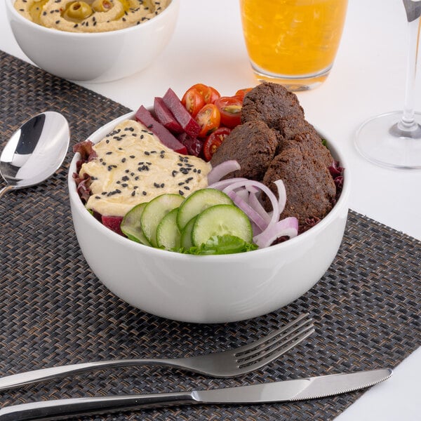 A white porcelain bowl filled with assorted fresh vegetables, falafel, and hummus.