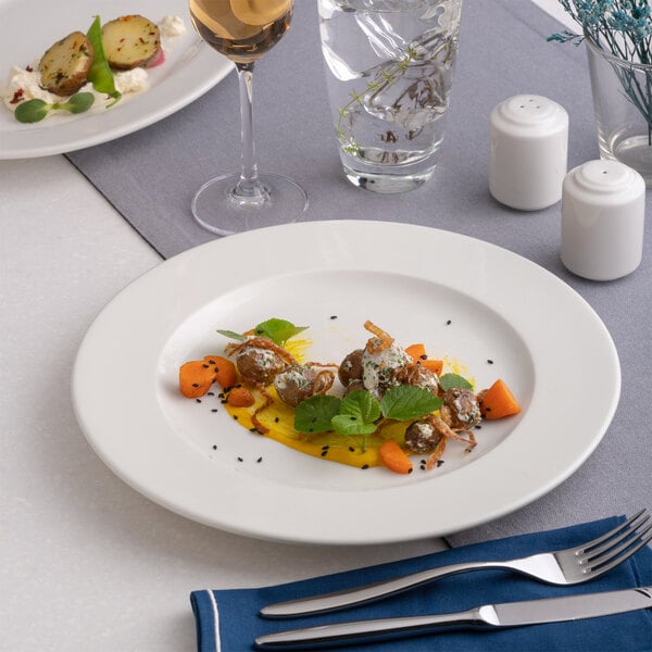 A round, wide rim, warm white porcelain plate with a flat center, shown as part of a table setting with food.