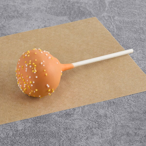 A pumpkin-flavored cake pop coated in orange icing with white and yellow sprinkles on a stick.