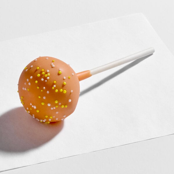 A pumpkin-flavored cake pop coated in orange icing and decorated with yellow and white sprinkles on a stick.
