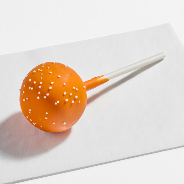 An orange cake pop with white sprinkles on a white stick, resting on a piece of white paper.