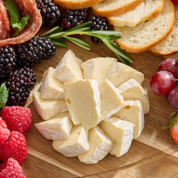 A wheel of Castello Brie cheese cut into wedges and arranged on a wooden board with assorted fruits, bread, and herbs.