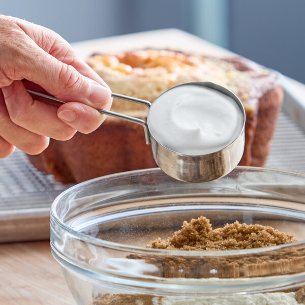 A metal measuring cup filled with white coconut yogurt alternative being held above a glass bowl containing brown sugar.