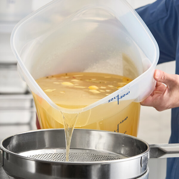 A large translucent round polypropylene food storage container with measurement markings being used to pour liquid through a strainer.