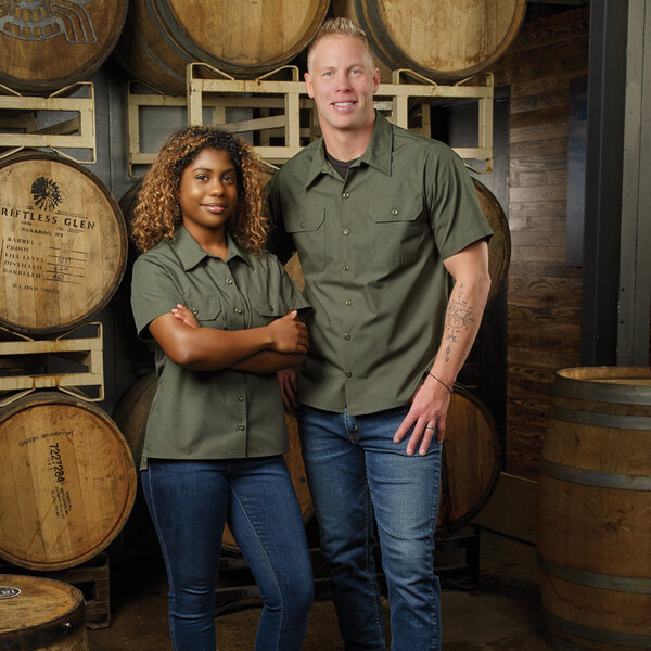 A woman wearing an olive short sleeve work shirt and jeans stands next to a man in a similar outfit in front of wooden barrels.