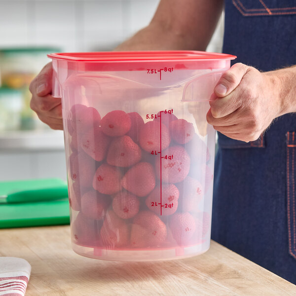 A translucent round polypropylene food storage container with a red lid, filled with strawberries and marked with measurement lines.