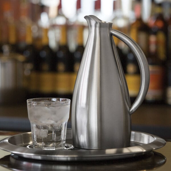 A brushed stainless steel pitcher and a glass of ice water on a metal tray.