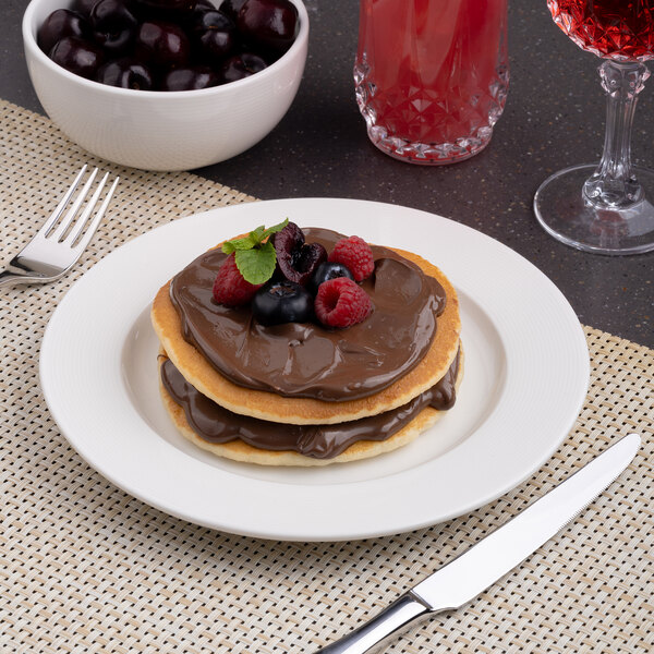 A white round embossed wide rim flat porcelain plate holding pancakes with chocolate spread and berries.