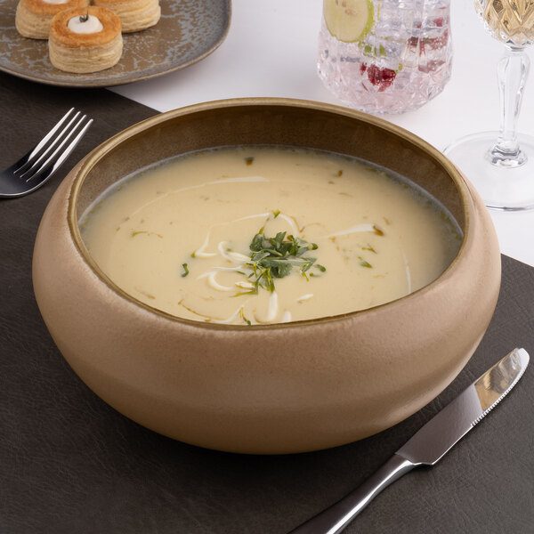 A large round gold and brown porcelain bowl filled with creamy soup, placed on a table setting.