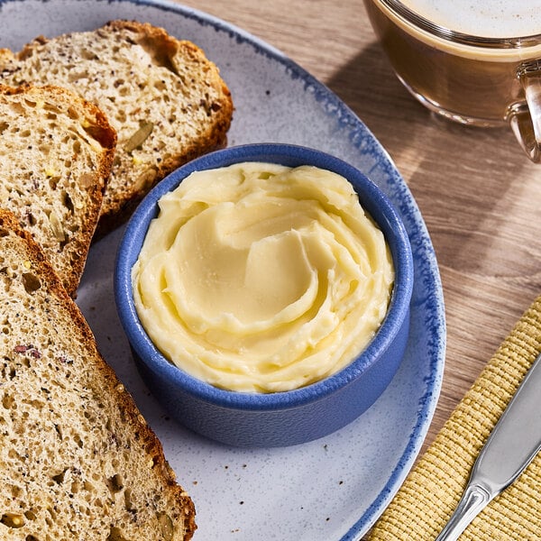 A blue ramekin filled with a swirl of European-style salted butter blend margarine, served on a plate with slices of multigrain bread.