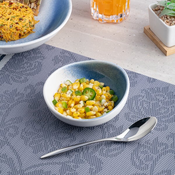 A small round blue and white ombre porcelain coupe bowl filled with corn salad, placed on a textured placemat with a spoon beside it.